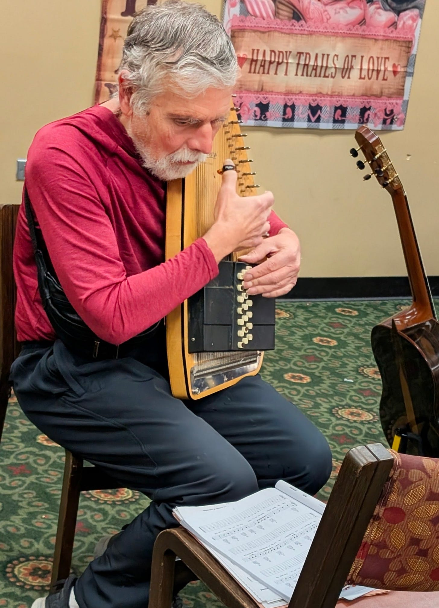 Silver-haired man in a red, long-sleeved athletic top plays an autoharp while looking intently at his music.