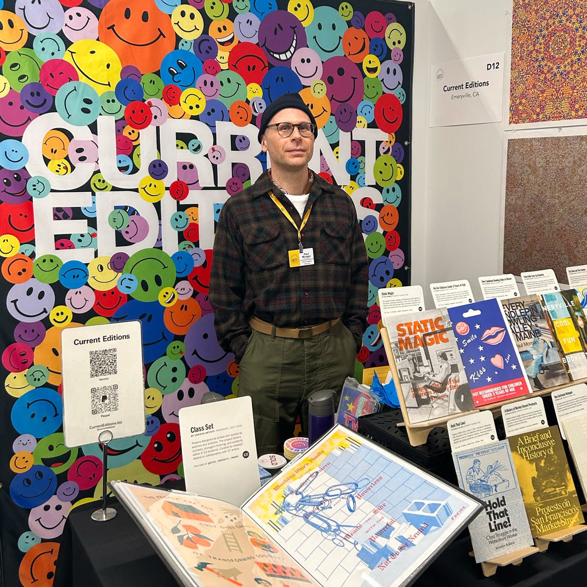A man wearing a beanie and glasses standing at the Current Editions booth at a book fair, with a colorful backdrop filled with multicolored smiley faces and bold white text reading "CURRENT EDITIONS." The booth, located at space D12, showcases a vibrant display of artist books, zines, and printed publications including titles like "Static Magic," "Hold That Line," and "A Brief and Incomprehensive History of Protests in San Francisco."