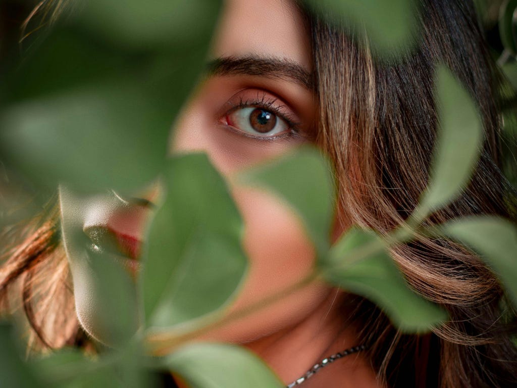 Close-up van een vrouwelijk gezicht dat gedeeltelijk achter groene bladeren verborgen is, met één oog zichtbaar. Close-up van een vrouwelijk gezicht dat gedeeltelijk achter groene bladeren verborgen is, met één oog zichtbaar.