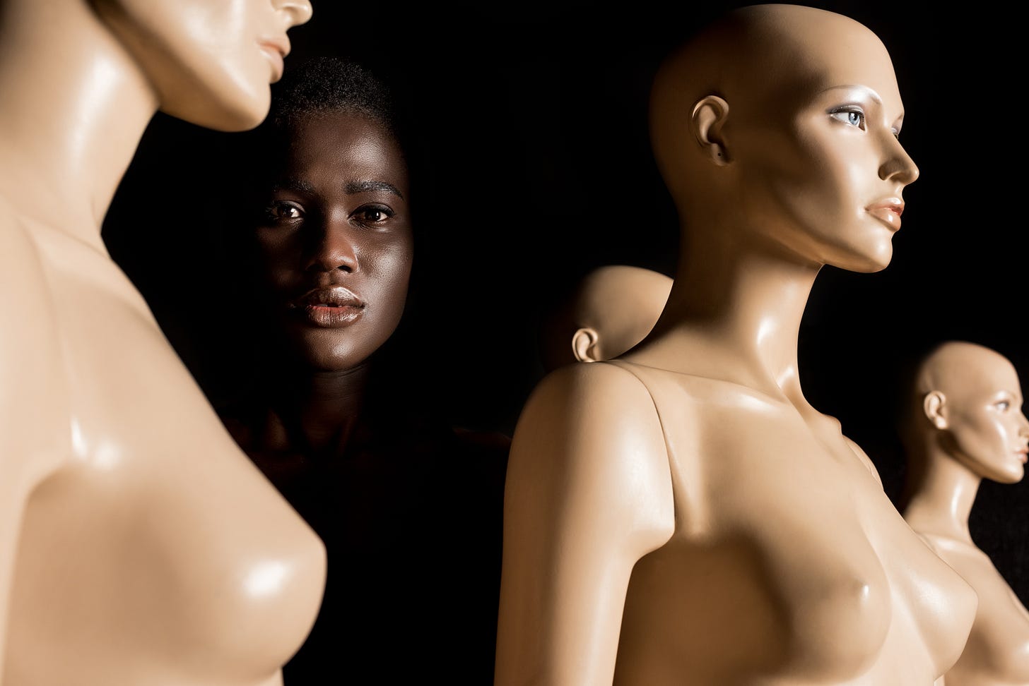 A Black woman stands in shadow among a group of nude mannequins, her real skin contrasting with the plastic bodies around her. A Black woman stands in shadow among a group of nude mannequins, her real skin contrasting with the plastic bodies around her.