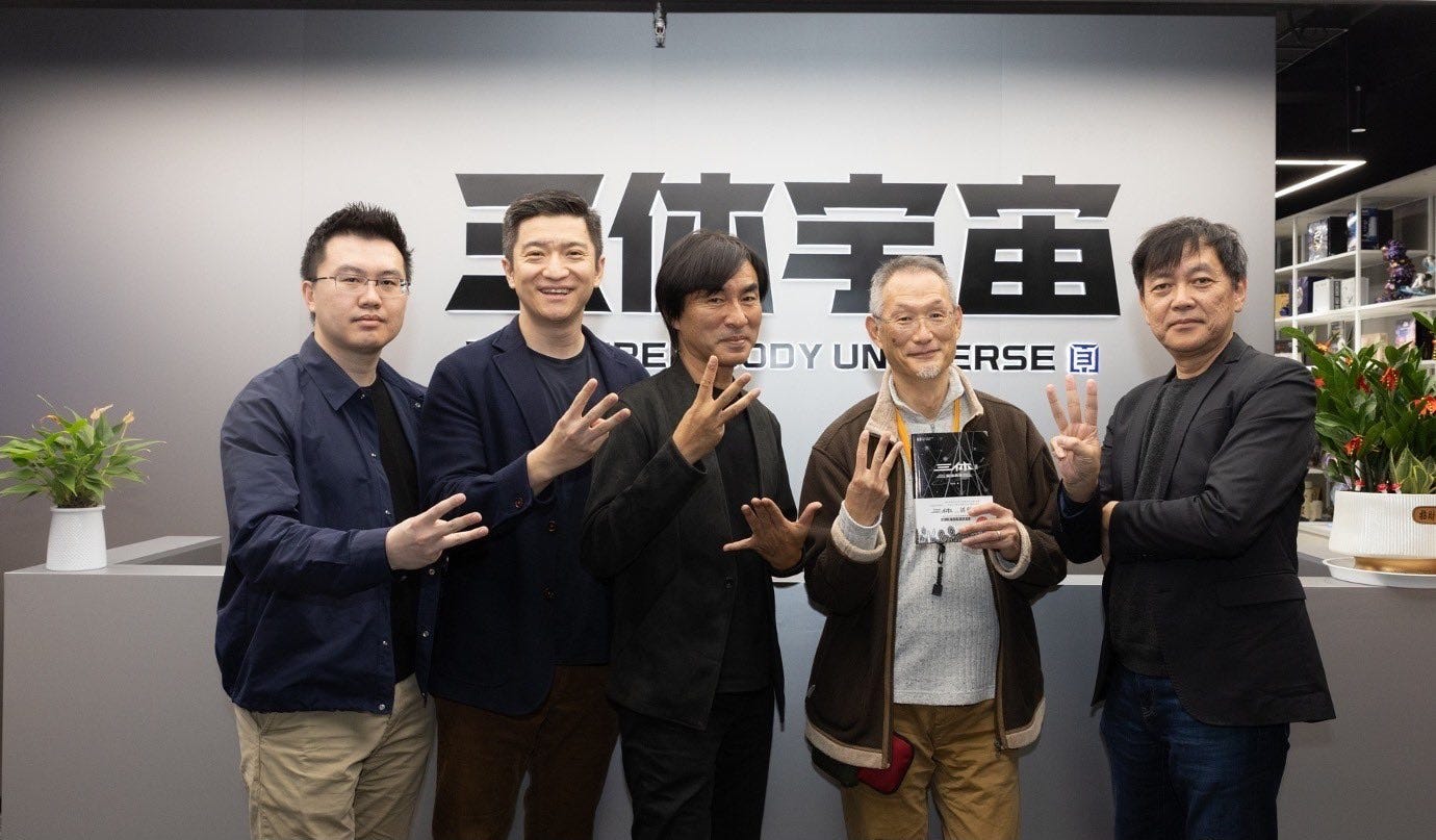 Photograph of five men standing next to each other in an office facing the camera. Each is holding up the three middle fingers of his right hand. Behind them are four Chinese characters and the words "The Three-Body Universe" displayed on the wall. Photograph of five men standing next to each other in an office facing the camera. Each is holding up the three middle fingers of his right hand. Behind them are four Chinese characters and the words "The Three-Body Universe" displayed on the wall.