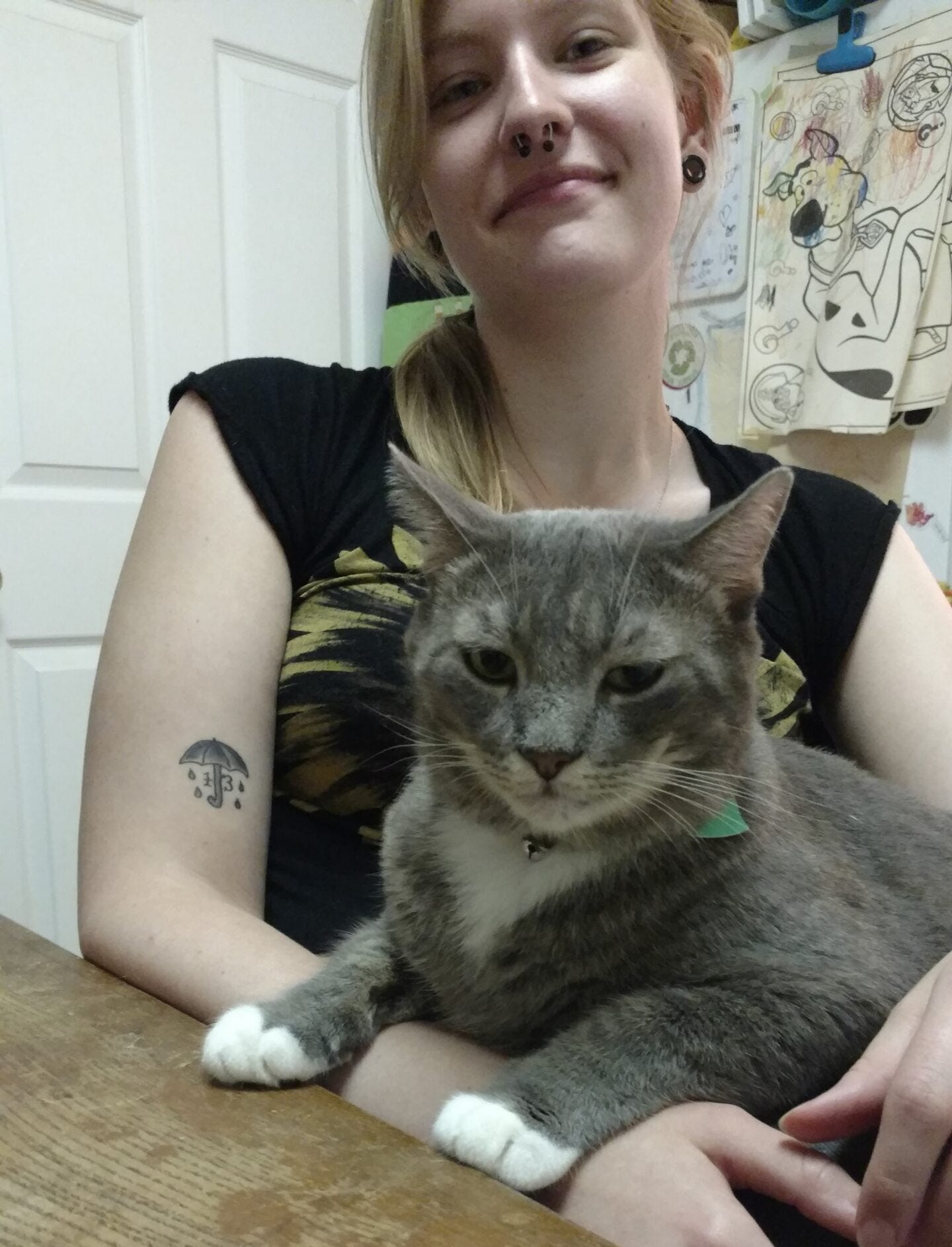 Savanna sitting with a grey cat in her lap at a brown wooden table in front of a fridge with colored illustrations on it. Savanna sitting with a grey cat in her lap at a brown wooden table in front of a fridge with colored illustrations on it.