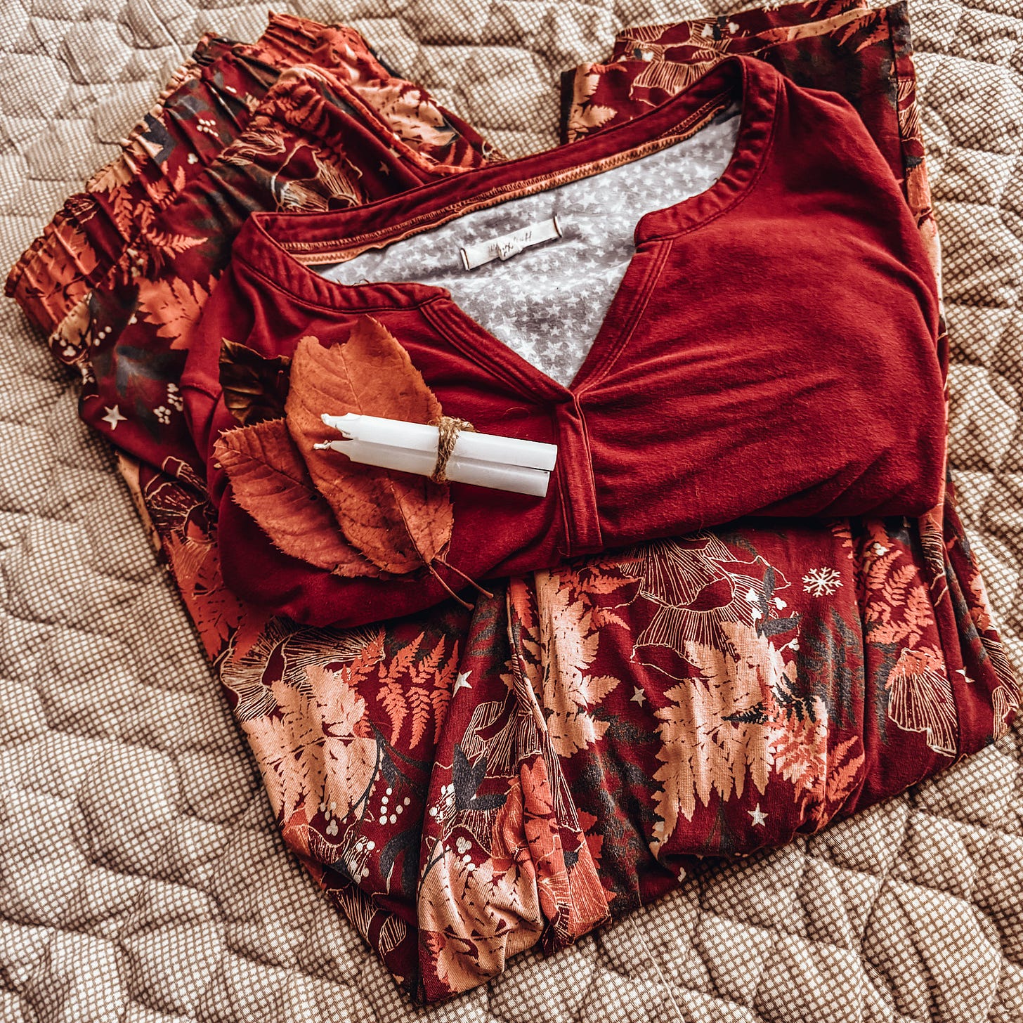 A set of pyjamas are shown on a light brown comforter. The pyjama top is a rustic red, with a pale grey inner with white stars. The trousers are red with florals in different autumnal colours. A trio of fallen autumn leaves, along with a bundle of three white candles, tied with twine, sit atop the pyjamas.