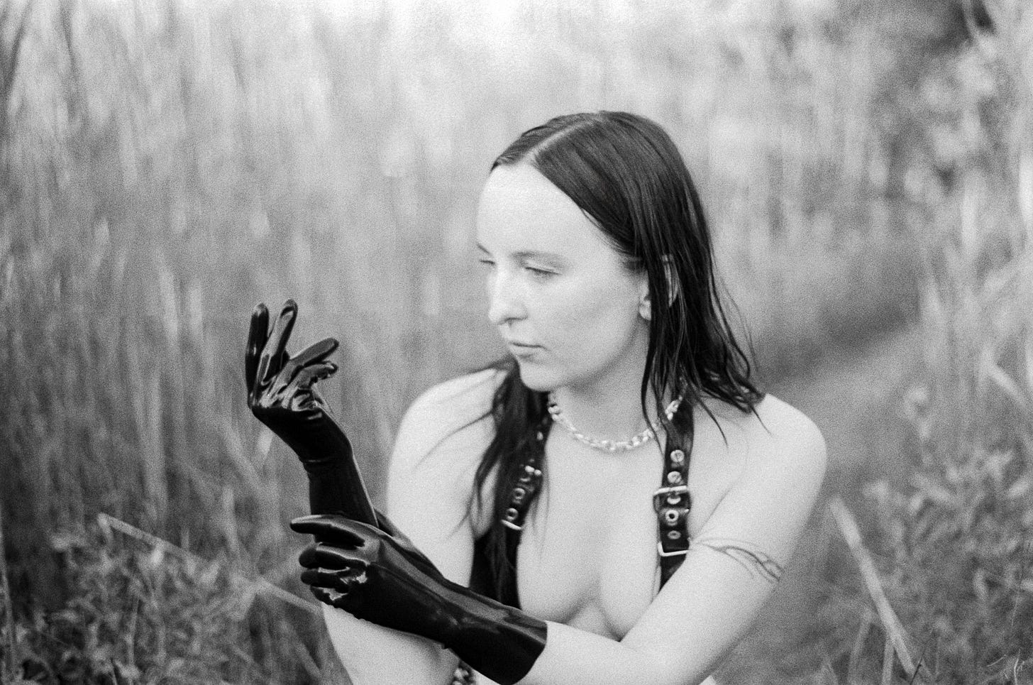 A black and white film portrait of Evyn Silver, a white femme with long dark hair, surrounded by tall beach grass. She wears black rubber gloves, a black rubber bikini with buckles, and a silver chain necklace with spikes. She is raising one hand in front of her face and smoothing out her glove with the other hand.