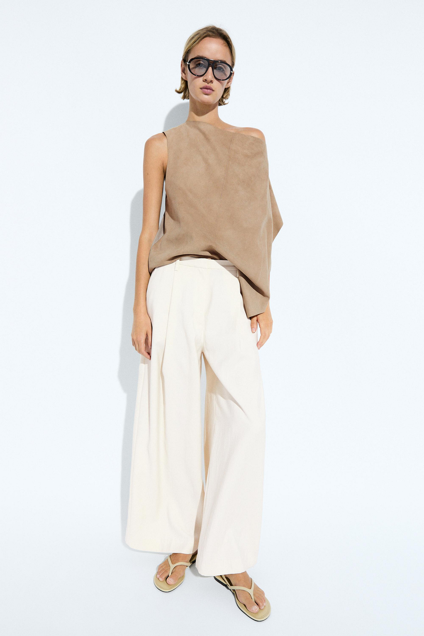 Ensemble composé d’un haut asymétrique sans manches couleur pierre avec détail drapé, une jupe midi claire, des chaussures plates foncées et des lunettes de soleil.
