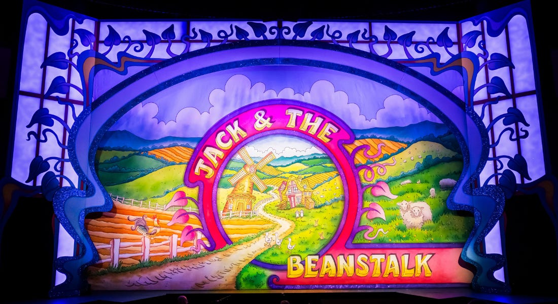 A vibrant, hand-painted cloth, depicting a countryside scene. A pink circle in the centre surrounds a windmill and farm. The words ‘Jack & The Beanstalk’ are in bold yellow lettering