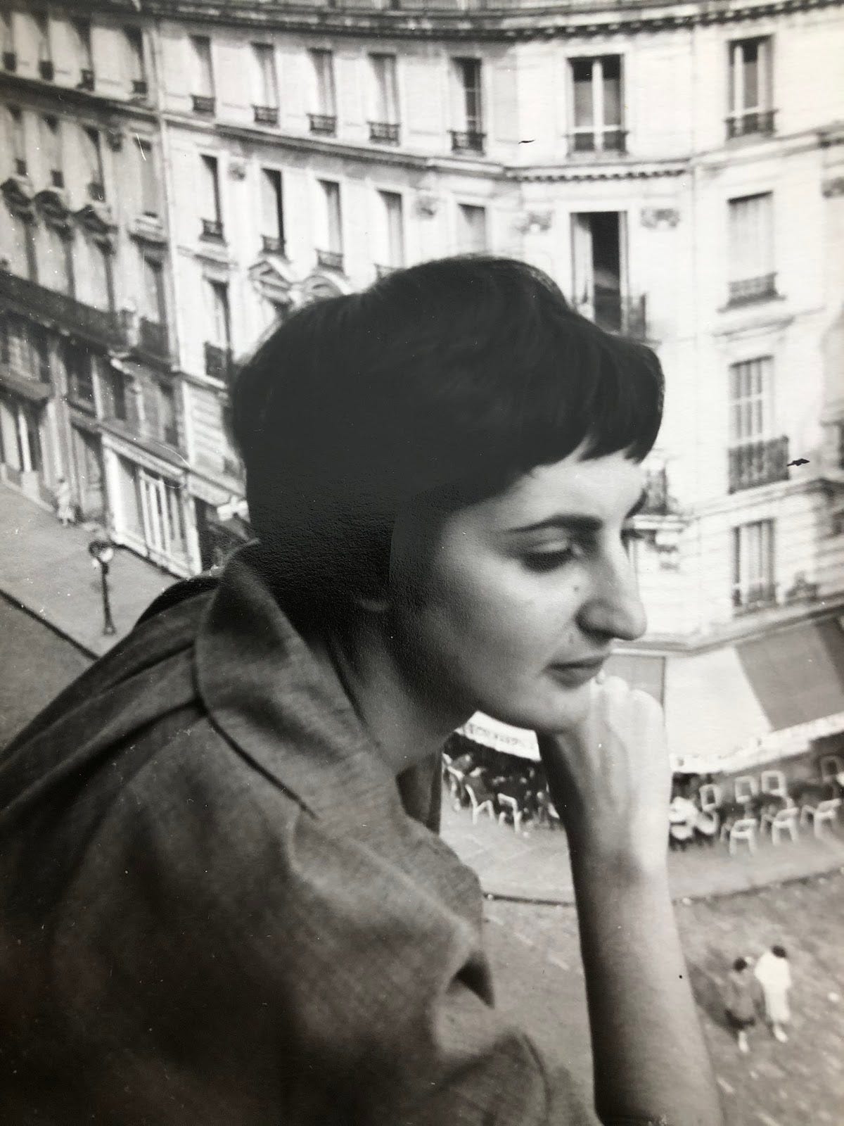 A three-quarter profile portrait of a woman with shoulders on a balcony high above a Paris street, her chin to the back of her hand, elbow on a railing.