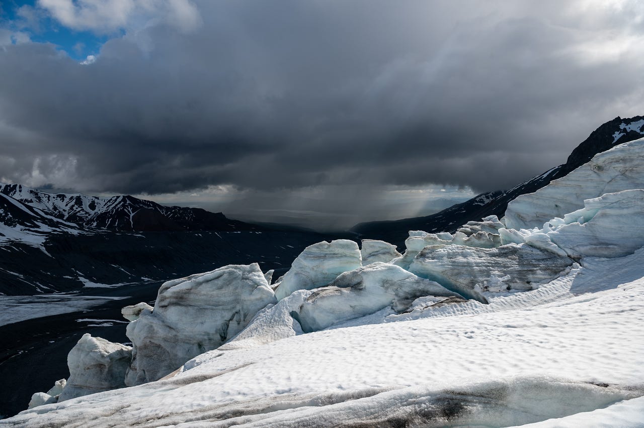 From the top of the icefall's snow-covered seracs, the view runs down a dark moraine valley to distant lowlands, where a grey column of rain pours from a huge storm cloud onto the plain around Summit Lake, the water just catching a strip of silver light. Snow-patched dark ridges frame the valley, with one corner of blue sky surviving at upper left.