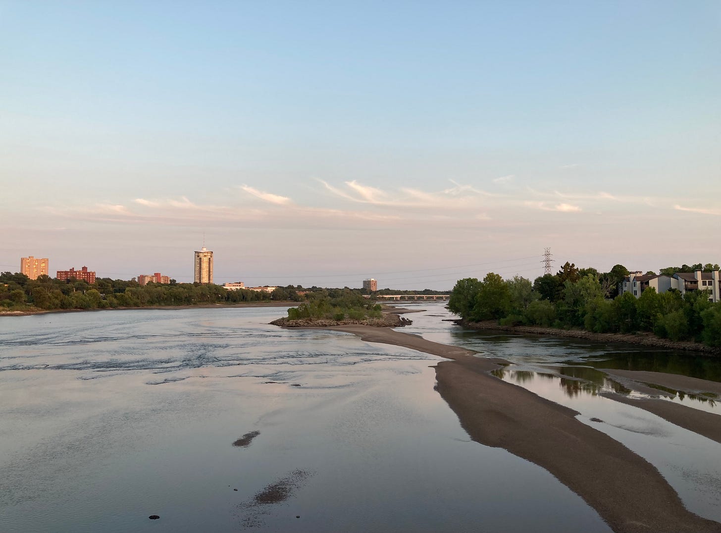 The photograph looks downstream over a wide, glassy Arkansas River, divided down one side by a bowing, terra cotta sandbar and vegetated island. The river reflects the fading pink and blue sky, streaked with delicate clouds just before dusk. In the distance, golden light illuminates the University Club Tower, Sophian Plaza and a few other buildings peeking out above the treetops along the east bank. The photograph looks downstream over a wide, glassy Arkansas River, divided down one side by a bowing, terra cotta sandbar and vegetated island. The river reflects the fading pink and blue sky, streaked with delicate clouds just before dusk. In the distance, golden light illuminates the University Club Tower, Sophian Plaza and a few other buildings peeking out above the treetops along the east bank.