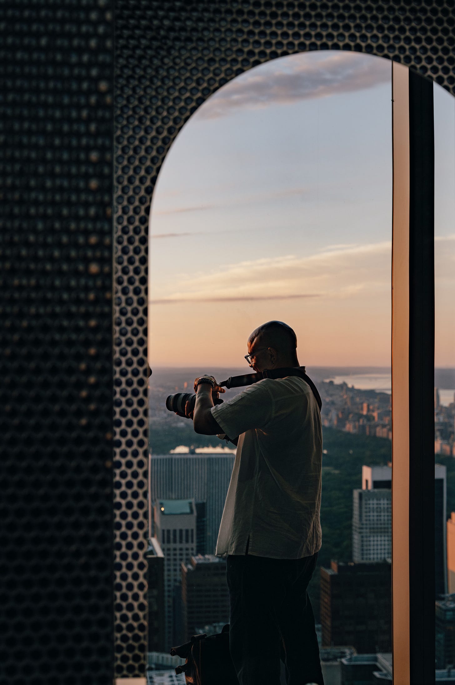 A man in a pale shirt photographing the sunrise through an arched opening in a dark perforated screen, the sky pink behind him. A man in a pale shirt photographing the sunrise through an arched opening in a dark perforated screen, the sky pink behind him.