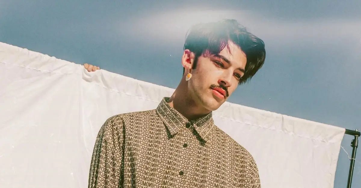 A photo of a white-presenting person with short brown hair that wears a patterned shirt and an earring in their left ear. They look down, out of the shot, and stand in front of a white sheet, mounted on a rail. A person's fingers are visible, at the top of the rail. The blue sky is visible in the background.
