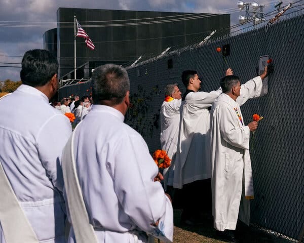 Religious leaders in white robes holding marigolds place them along a high fence topped with barbed wire. 