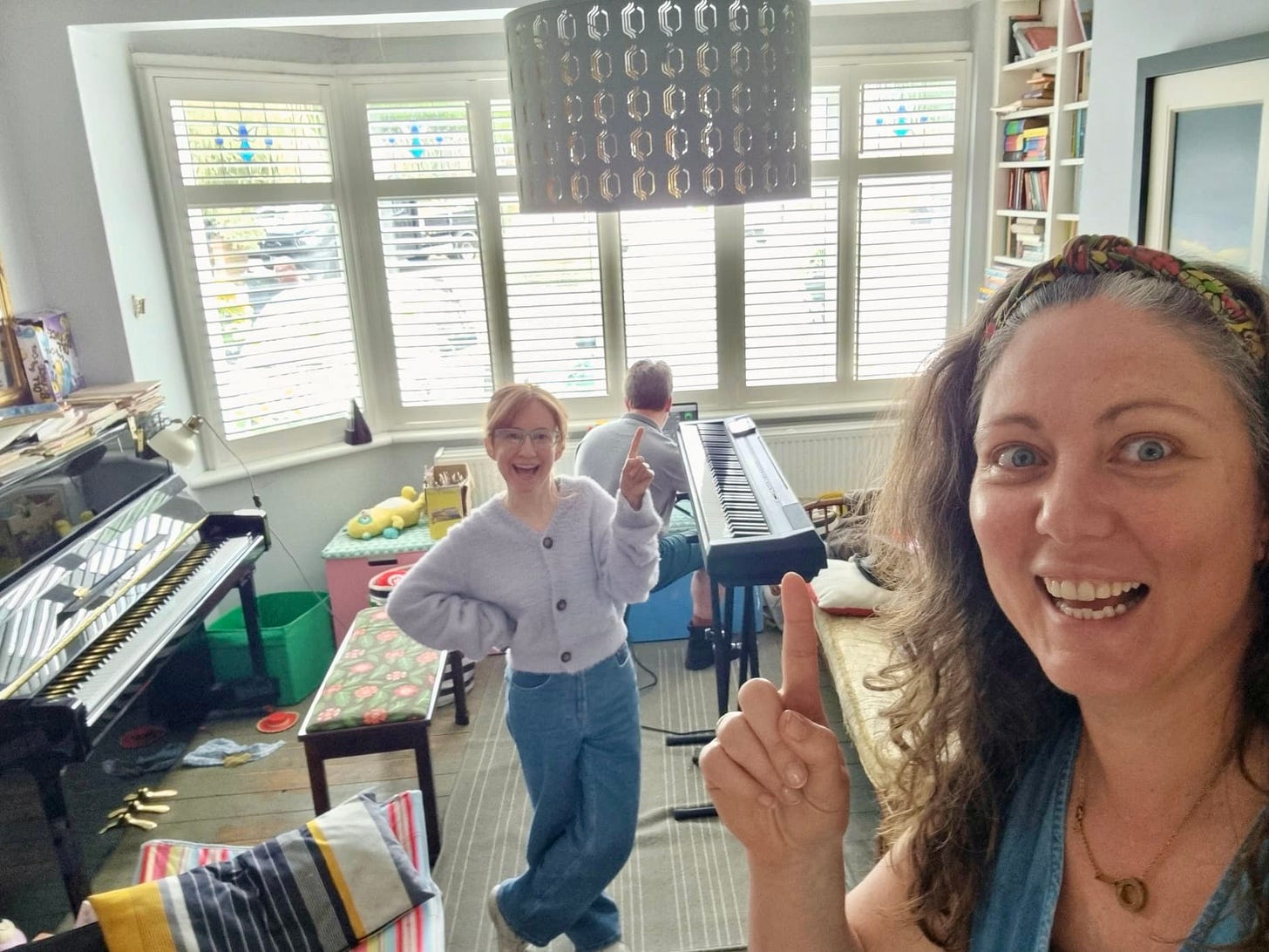 Two women point jauntily upwards smiling at the camera. They are in a front room with a keyboard and piano.  A man crouches by a laptop with his back to us