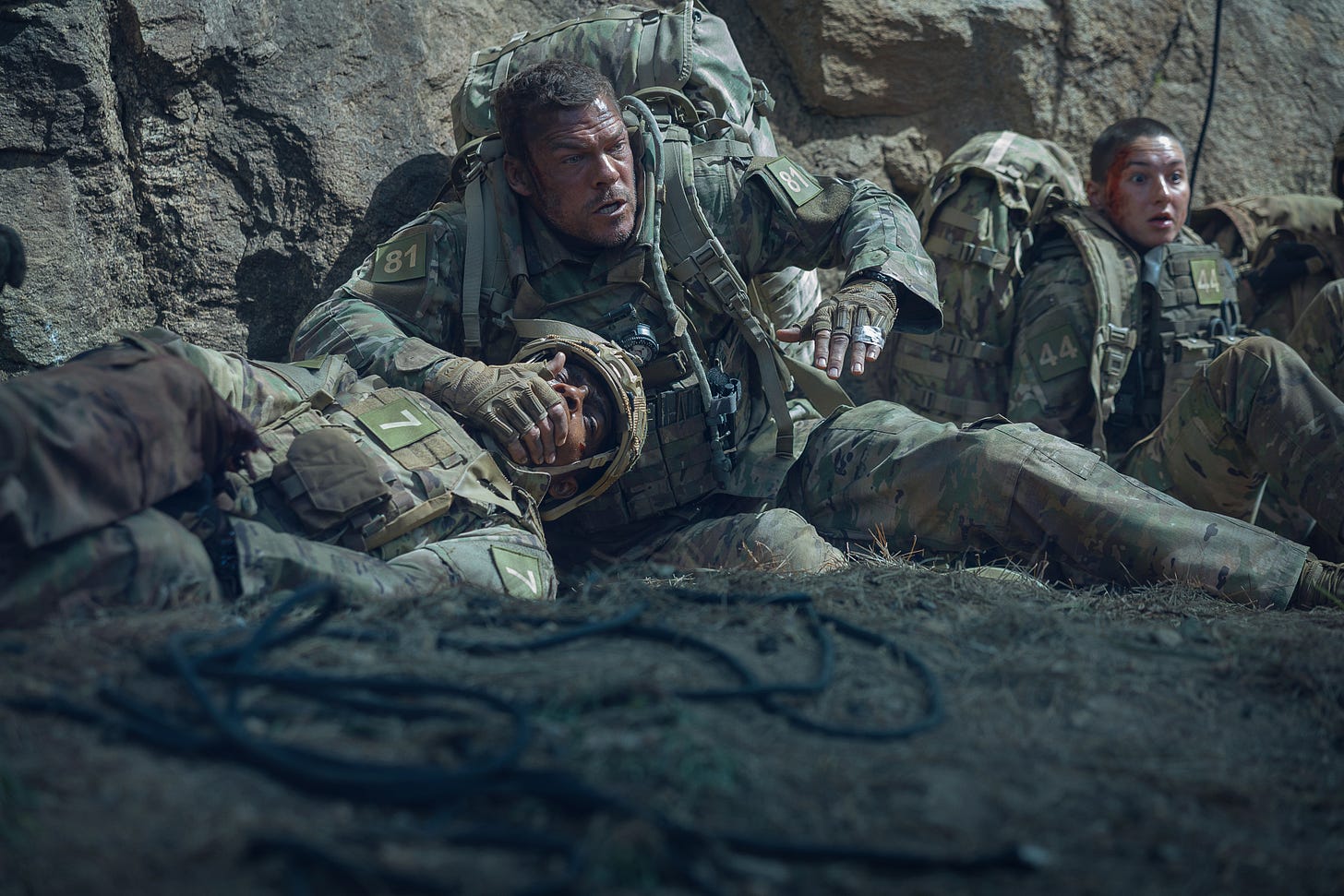 Ritchson as 81, hides next to a cliff wall, holding his hand over the mouth of prone soldier. 