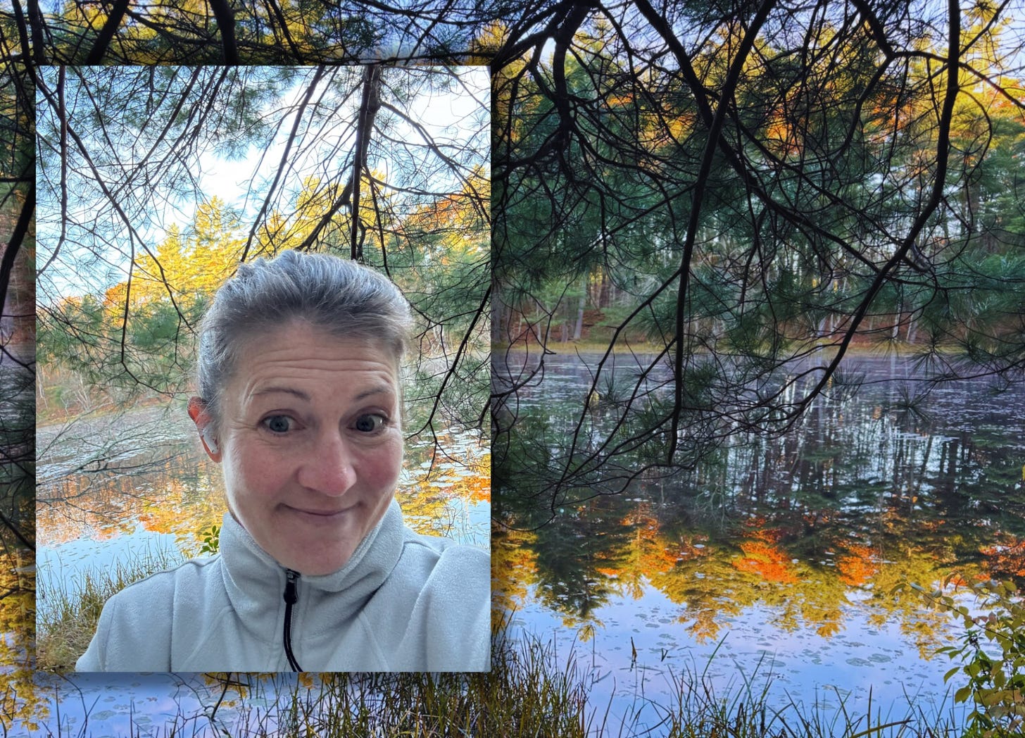 I’m talking a selfie outdoors near a calm pond surrounded by trees in autumn. The pond reflects the colorful fall foliage and branches above, creating a mirrored effect on the water. 