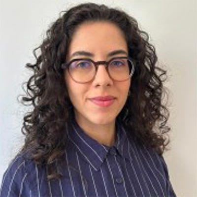 Jovem de cabelos escuros, volumosos e com cachos bem definidos. Ela utiliza óculos de armação arredondada em tom marrom. Ela veste uma camisa social de cor azul-escura com padronagem de risca de giz fina em branco. Jovem de cabelos escuros, volumosos e com cachos bem definidos. Ela utiliza óculos de armação arredondada em tom marrom. Ela veste uma camisa social de cor azul-escura com padronagem de risca de giz fina em branco.