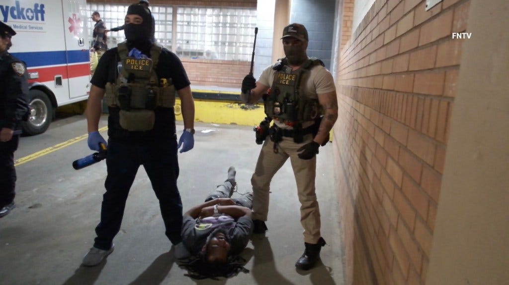 Two ICE officers, dressed in tactical gear and face coverings, stand over a man lying on the ground, with an ambulance visible in the background. Two ICE officers, dressed in tactical gear and face coverings, stand over a man lying on the ground, with an ambulance visible in the background.