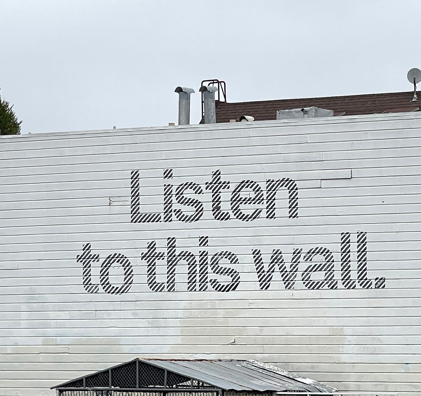 large white-painted wall with bold black striped letters spelling “Listen to this wall.” Rooftop vents, antennas, and a satellite dish rise behind the building, and part of a slanted metal roof is visible at the bottom of the frame.
