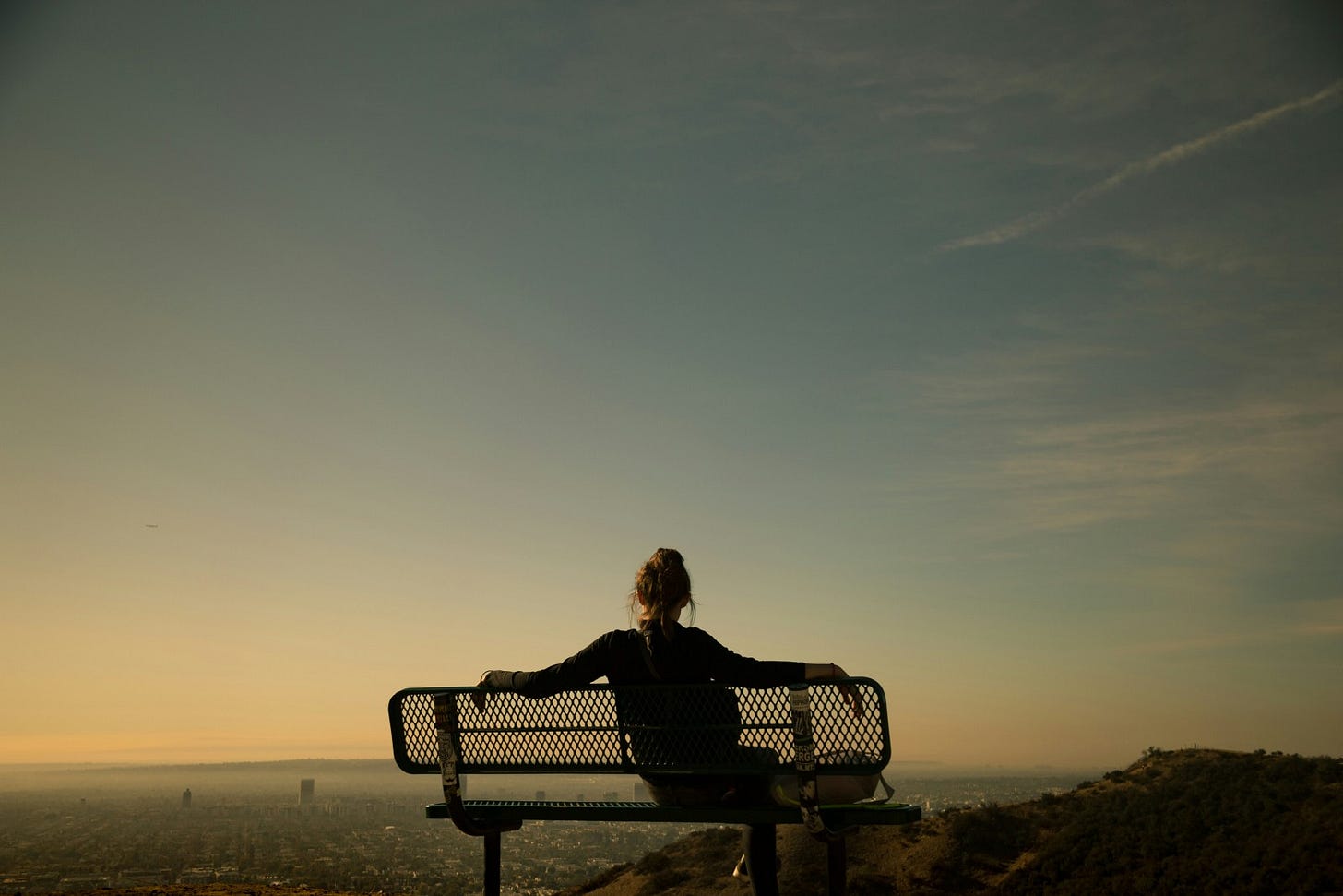 Seen from behind, a person sits on a bench with outstretched arms, contemplating the sprawling city in the distance. The image evokes a powerful sense of personal reflection and surrender against a serene, misty landscape.