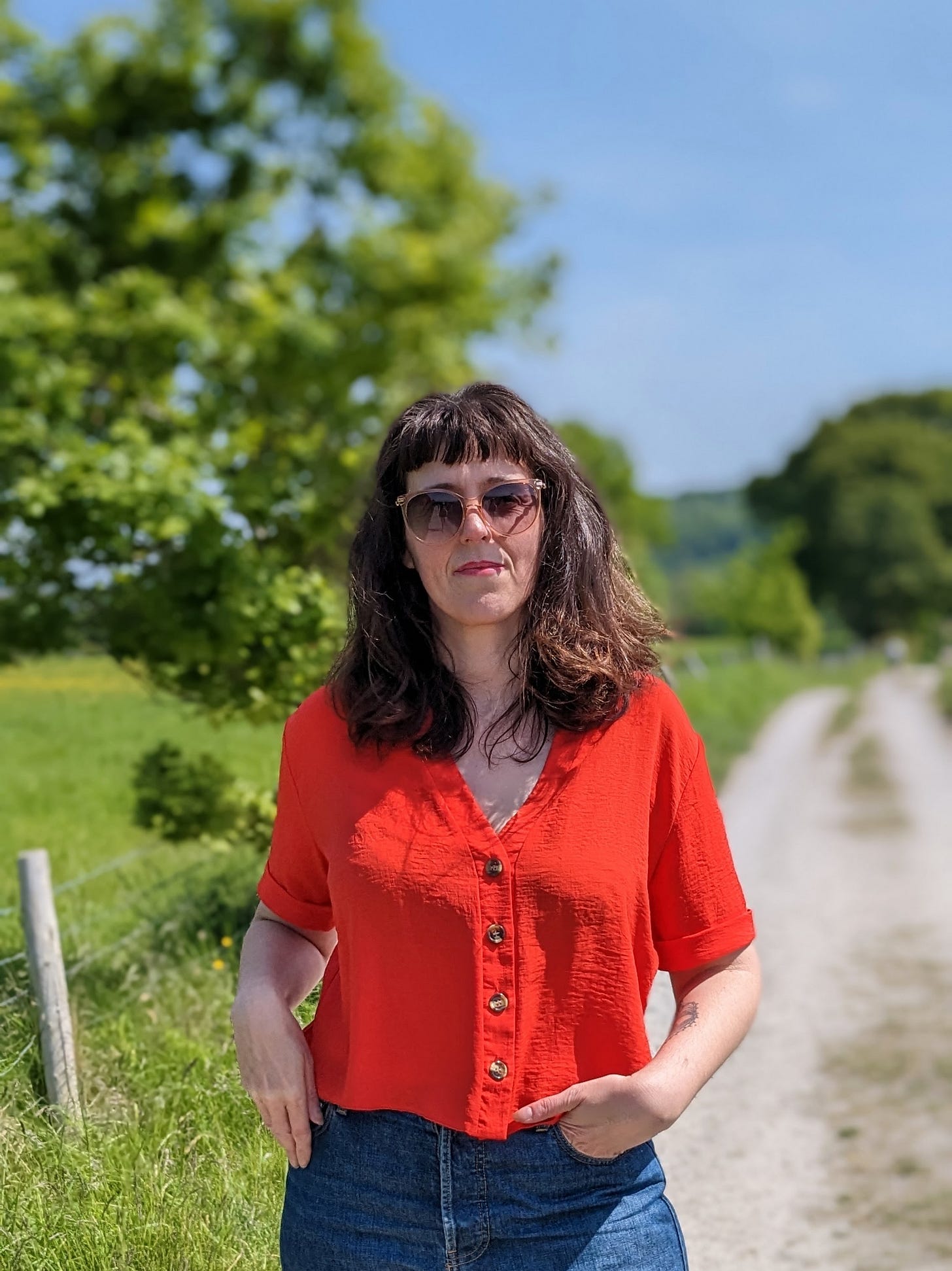 susan earlam stood facing the camera on a footpath. she wears sunglasses and a red top, it is a sunny day