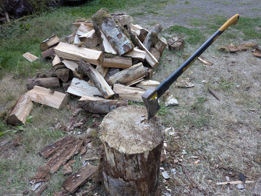 An ax with its handle sticking out, the blade stuck into a round chopping block.  A pile of split firewood sits in the background.