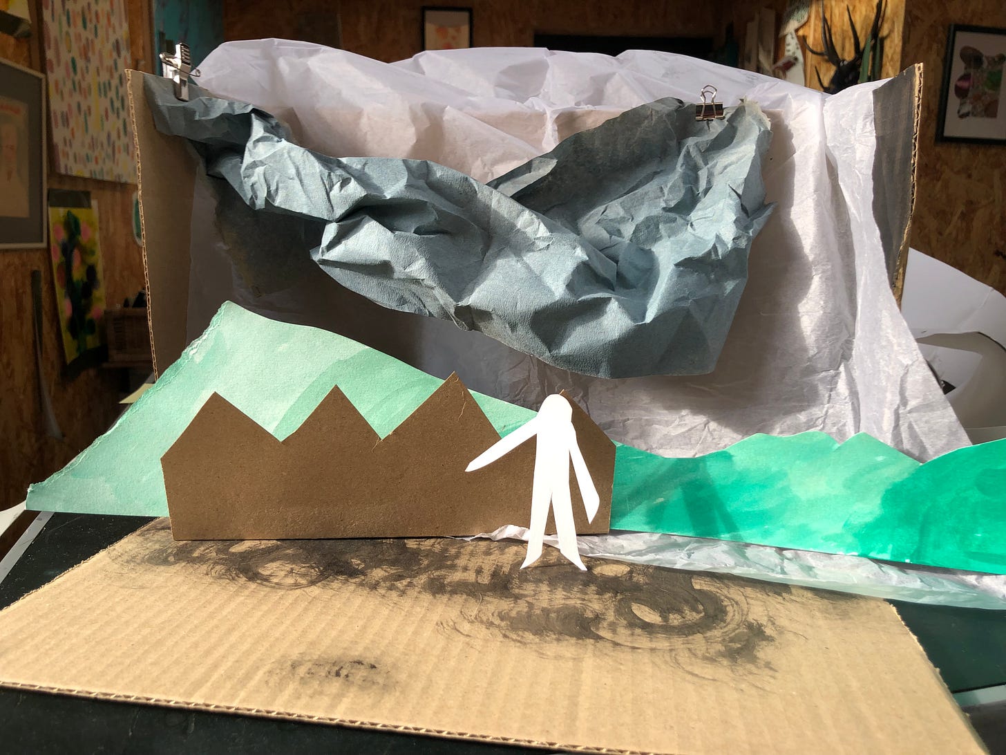 A paper figure stands in front of cut-out houses, hills and a crumpled paper sky, in a small scale set. 