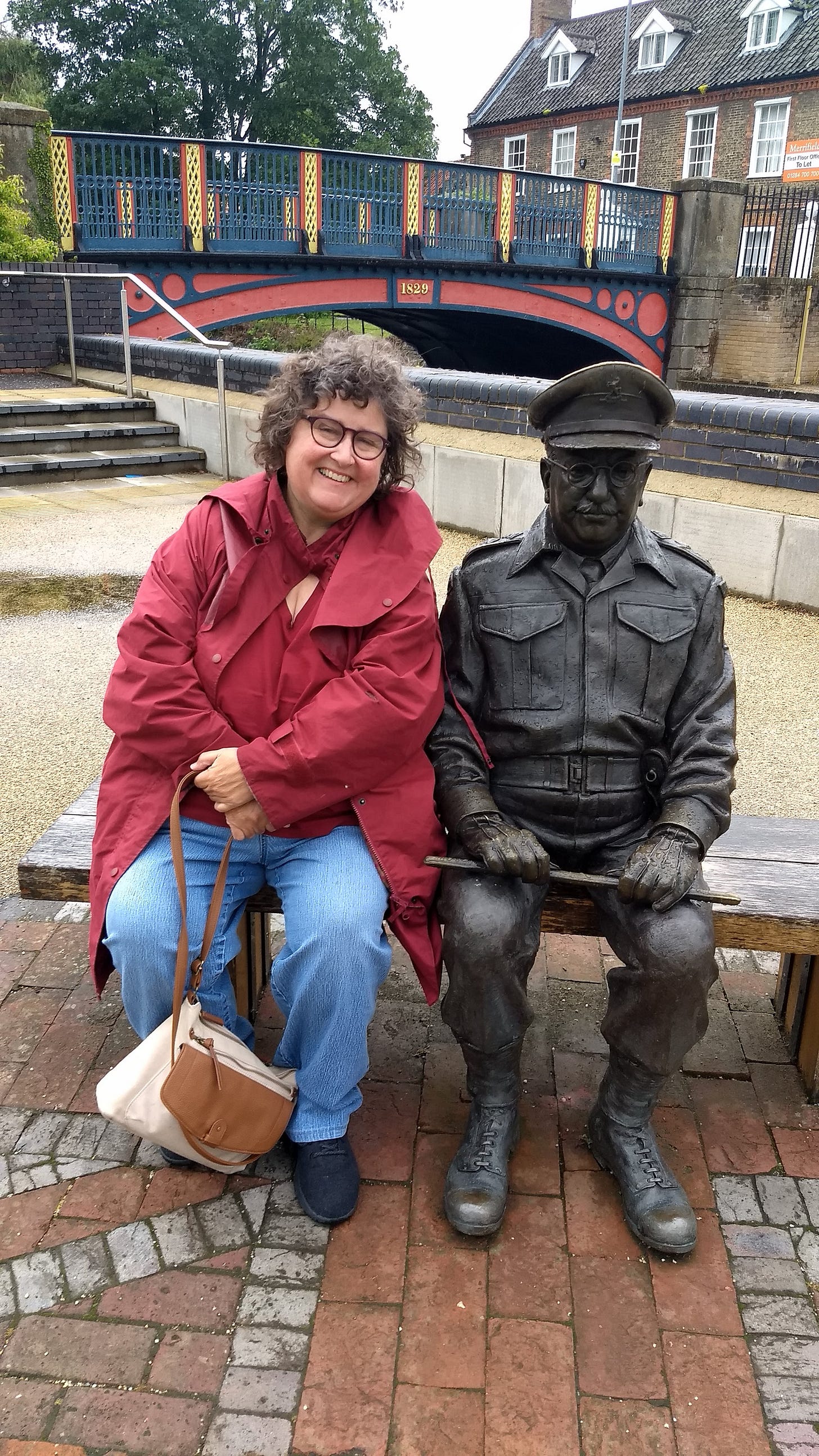 Annette on bench in rainjacket with statue of man in military uniform, sitting on bench