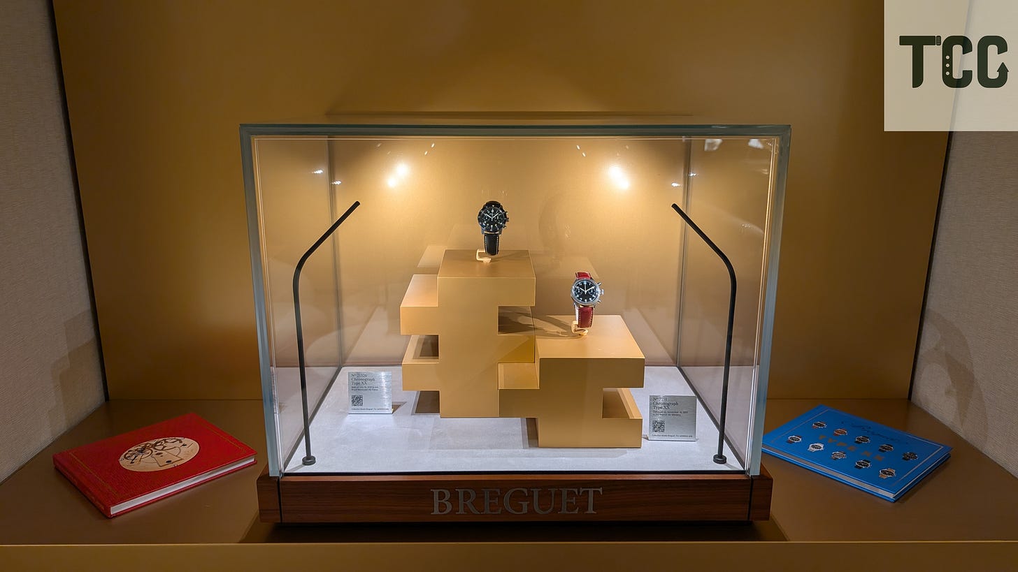 Glass display case with two Breguet pilot watches on golden pedestals, with a red book on the left and a blue booklet on the right.