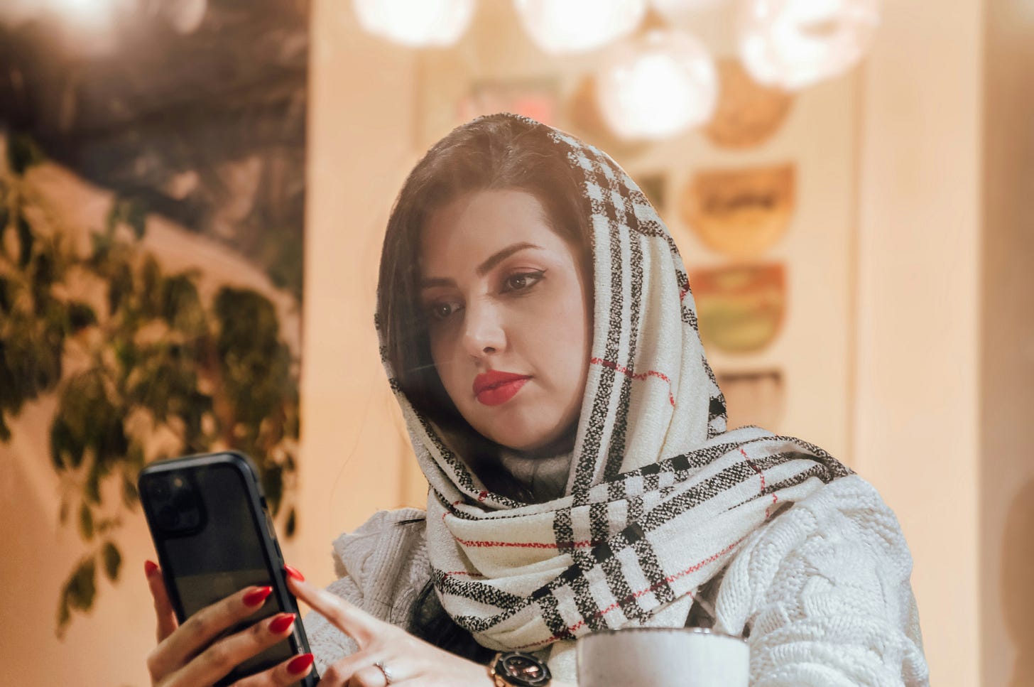 A woman with a plaid scarf wrapped around her head is sitting at an indoor café, looking at her phone.