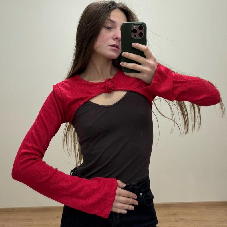 May include: A red bolero top with long sleeves and a keyhole detail at the chest. The top is worn over a black tank top. The sleeves are flared at the wrists. The person is holding a phone.