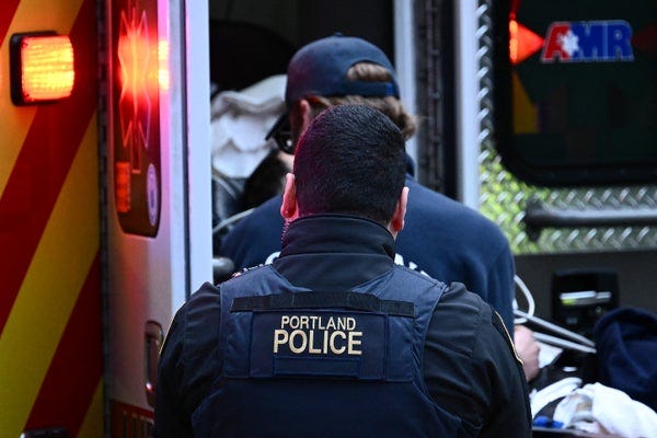 Photograph of the back of a Portland police officer as they watch an EMT help a patient inside an ambulance in Portland, Oregon Photograph of the back of a Portland police officer as they watch an EMT help a patient inside an ambulance in Portland, Oregon