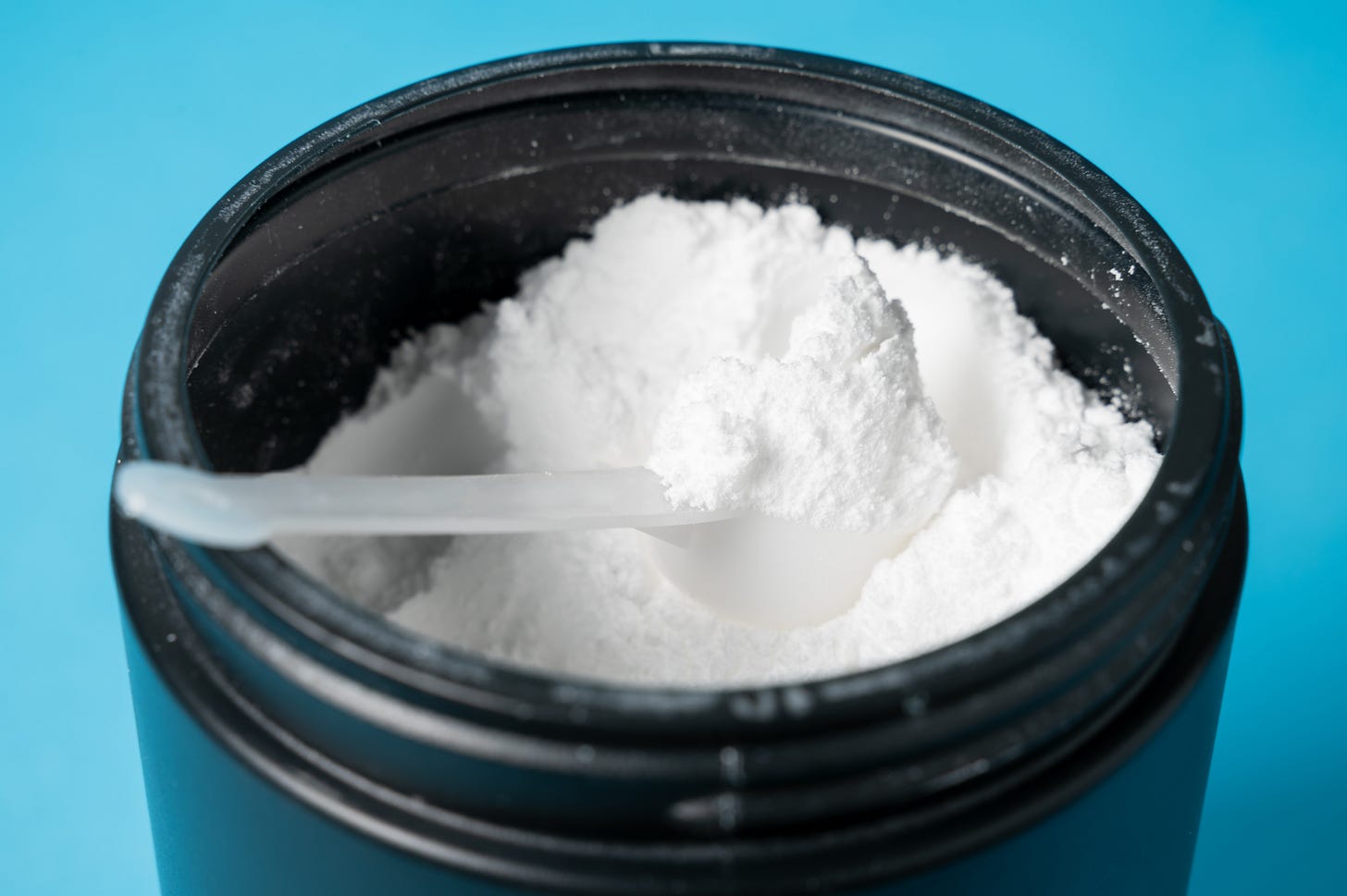 Close up of open canister of white powder supplement with a scoop in it.