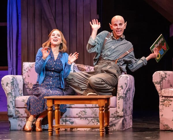 A man with pointy bat ears and a woman, seated together in a living room gesture giddily. A man with pointy bat ears and a woman, seated together in a living room gesture giddily.