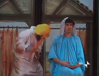 In una scena interna con una finestra sullo sfondo, due persone con costumi teatrali improvvisati. A sinistra, una persona in una veste chiara con in testa una sorta di berretto o turbante giallo si piega in avanti. A destra, una persona è seduta, indossa una veste azzurra e ha il capo coperto da un drappo bianco, con un'espressione neutra. Si intravede il logo "RAI" in basso a destra. In sovrimpressione la scritta bianca ANNUNCIAZIONE! ANNUNCIAZIONE!