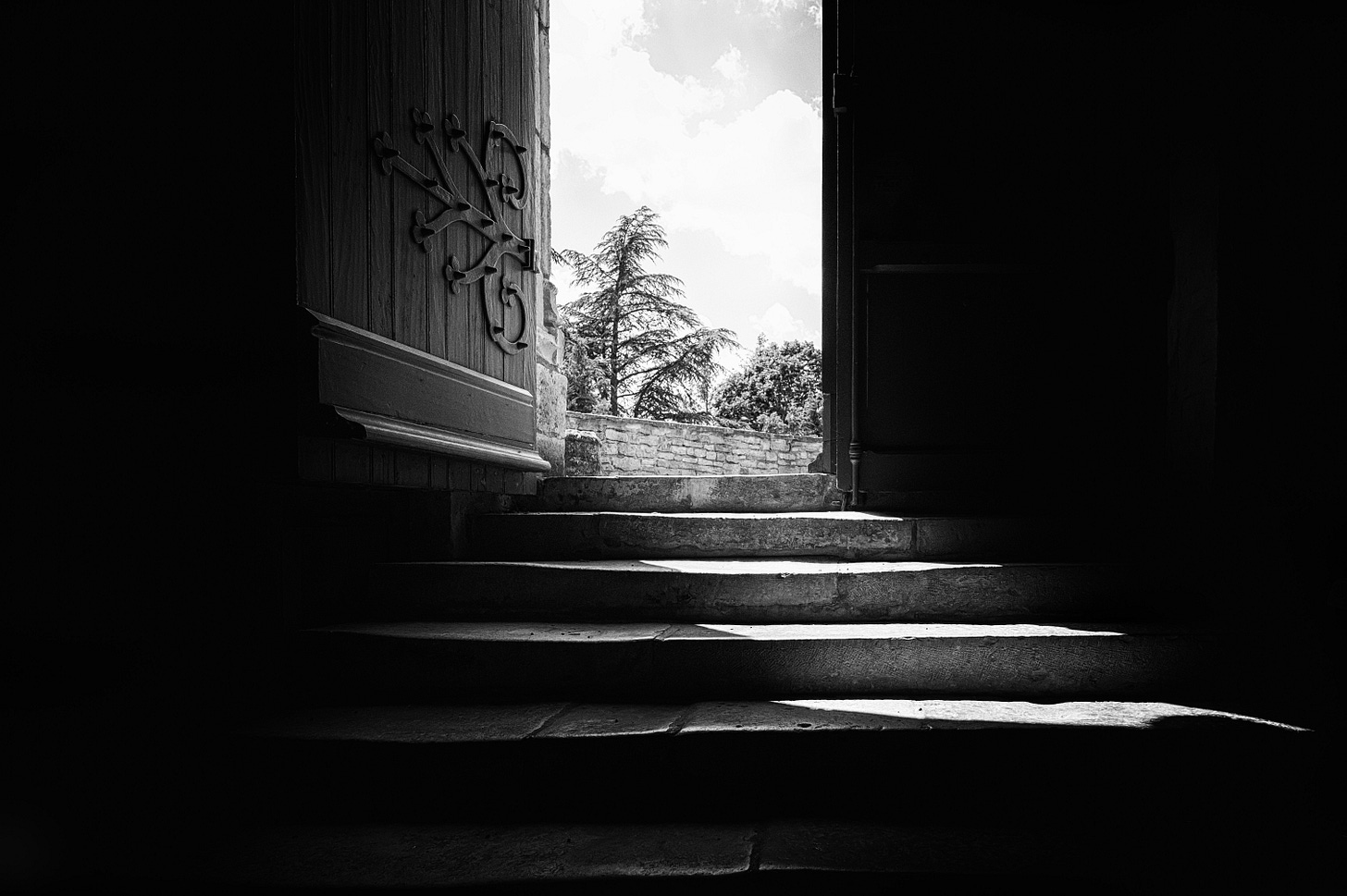 Black and white photo of a door open at the top of a few stone steps. The sky, trees, and a rockwall are in view of the open door.
