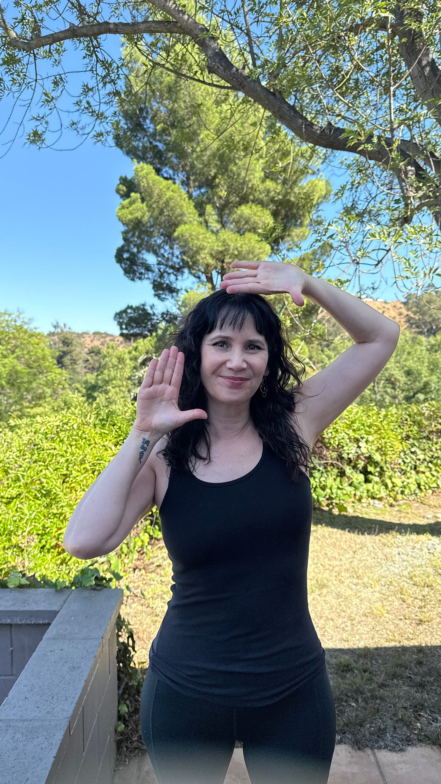 Laverne standing outside in a black tank top and black yoga pants. She is smiling while framing her face with her hands.