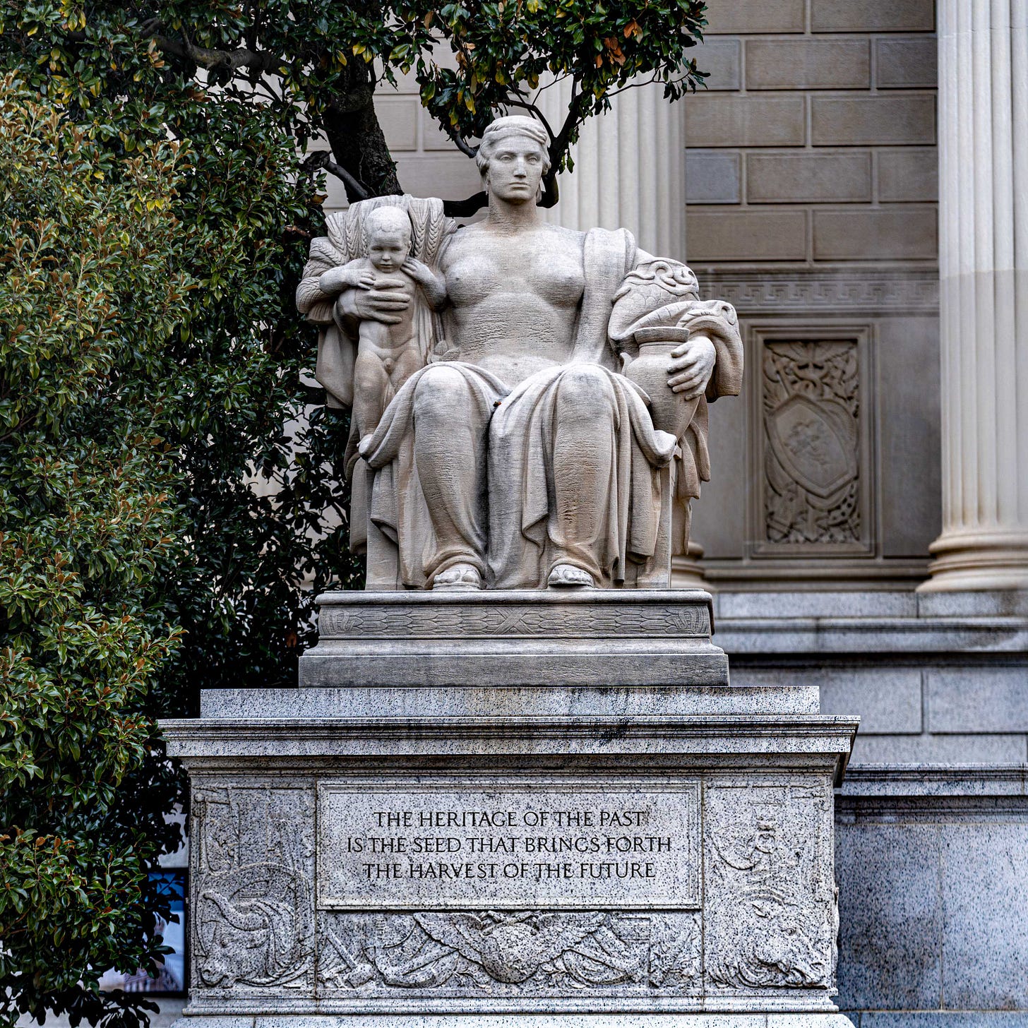‘Heritage’ in front of the United States National Archives in Washington, D.C., holding an infant in a sheaf of wheat in one arm, and a snake-topped urn in the other. The figure is seated on a Levantine throne, resting on a pedestal adorned with a winged solar disk, and the inscription: ”THE HERITAGE OF THE PAST IS THE SEED THAT BRINGS FORTH THE HARVEST OF THE FUTURE.” Photo by Peter Duke ©2026 - All Rights Reserved.