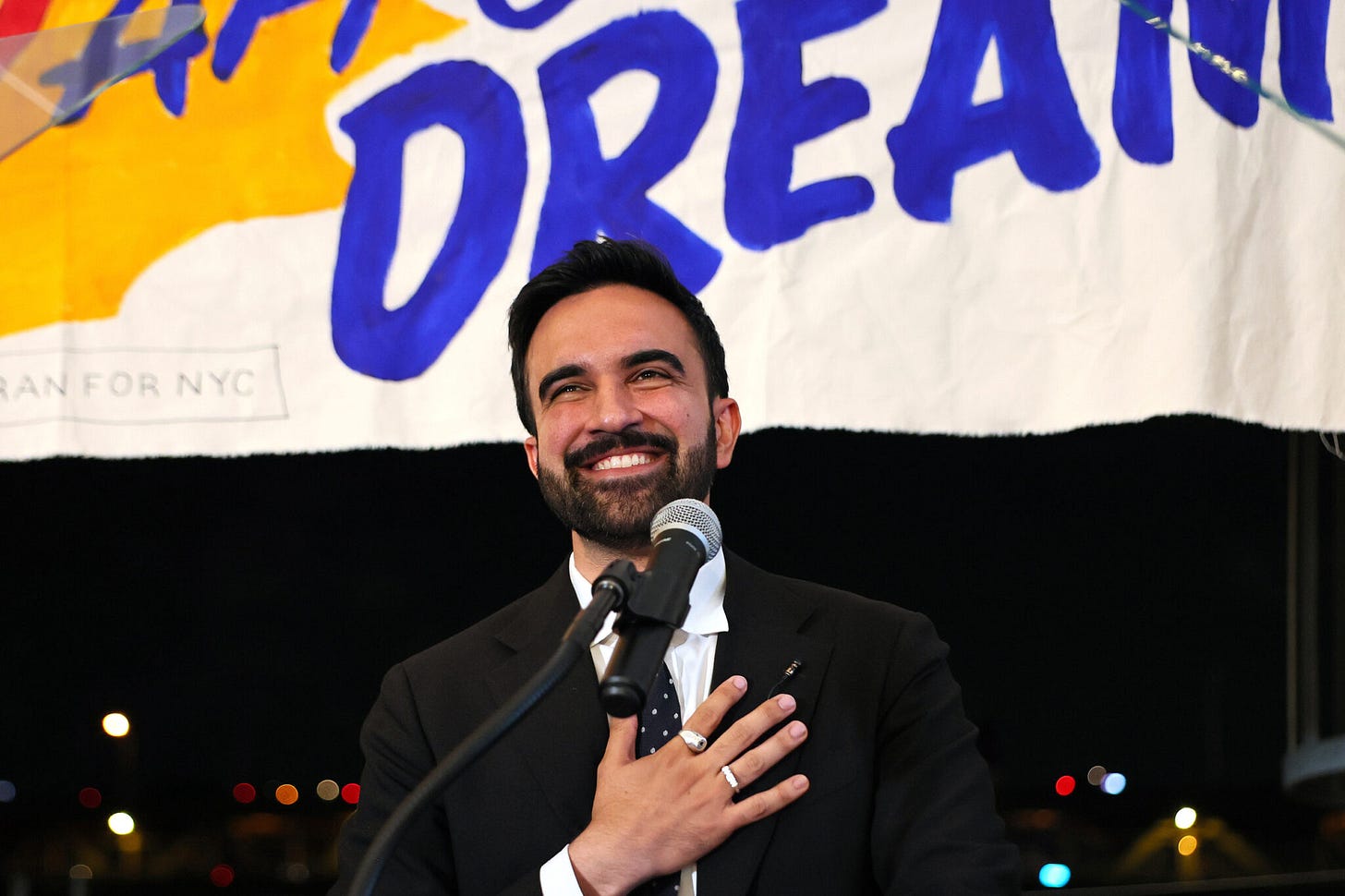 Zohran has dark hair and beard, he's smiling and has his hand on his chest. He is in front of a mic, with the backdrop of a poster which is obscured. 