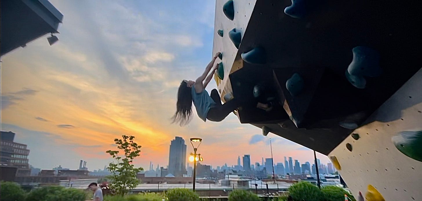 photo of a woman climbing an outdoor rock wall at sunset with a city skyline in the distance
