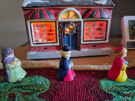 Small figures from a nativity scene in front of a model bakery and a DVD player. A small pottery sheep is sitting on top of a barn.