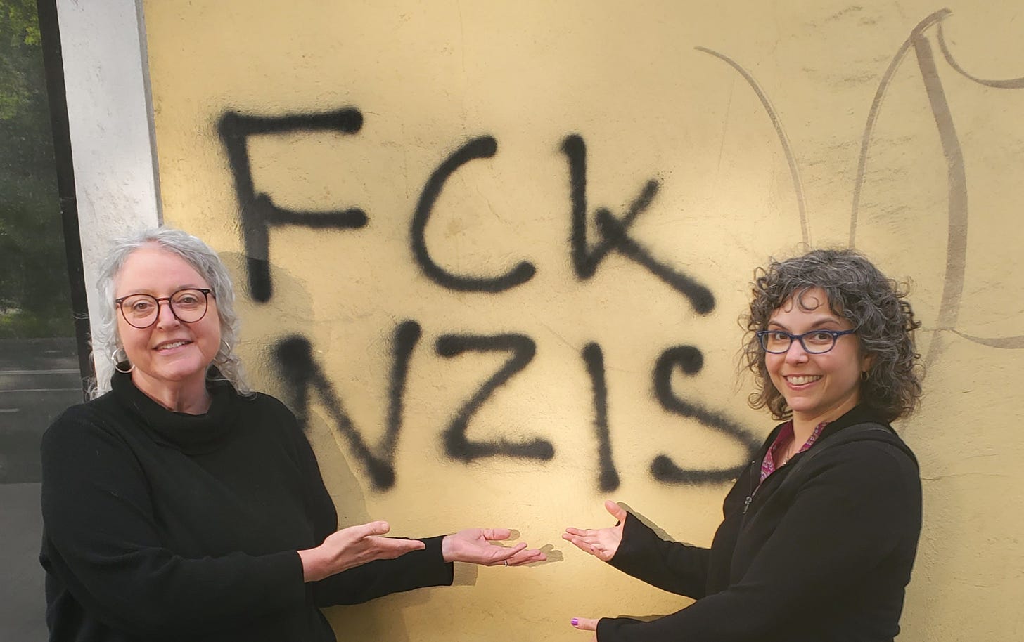 Two Jewish women in black tops, each wearing glasses, stand in front of graffiti that reads FCK NZIS