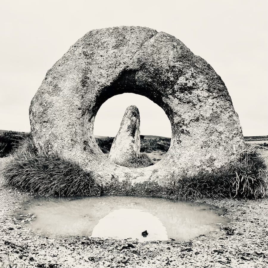 A black and white photograph of a granite squat stone with a perfect hole in the centre. Through it, another standing stone is framed.