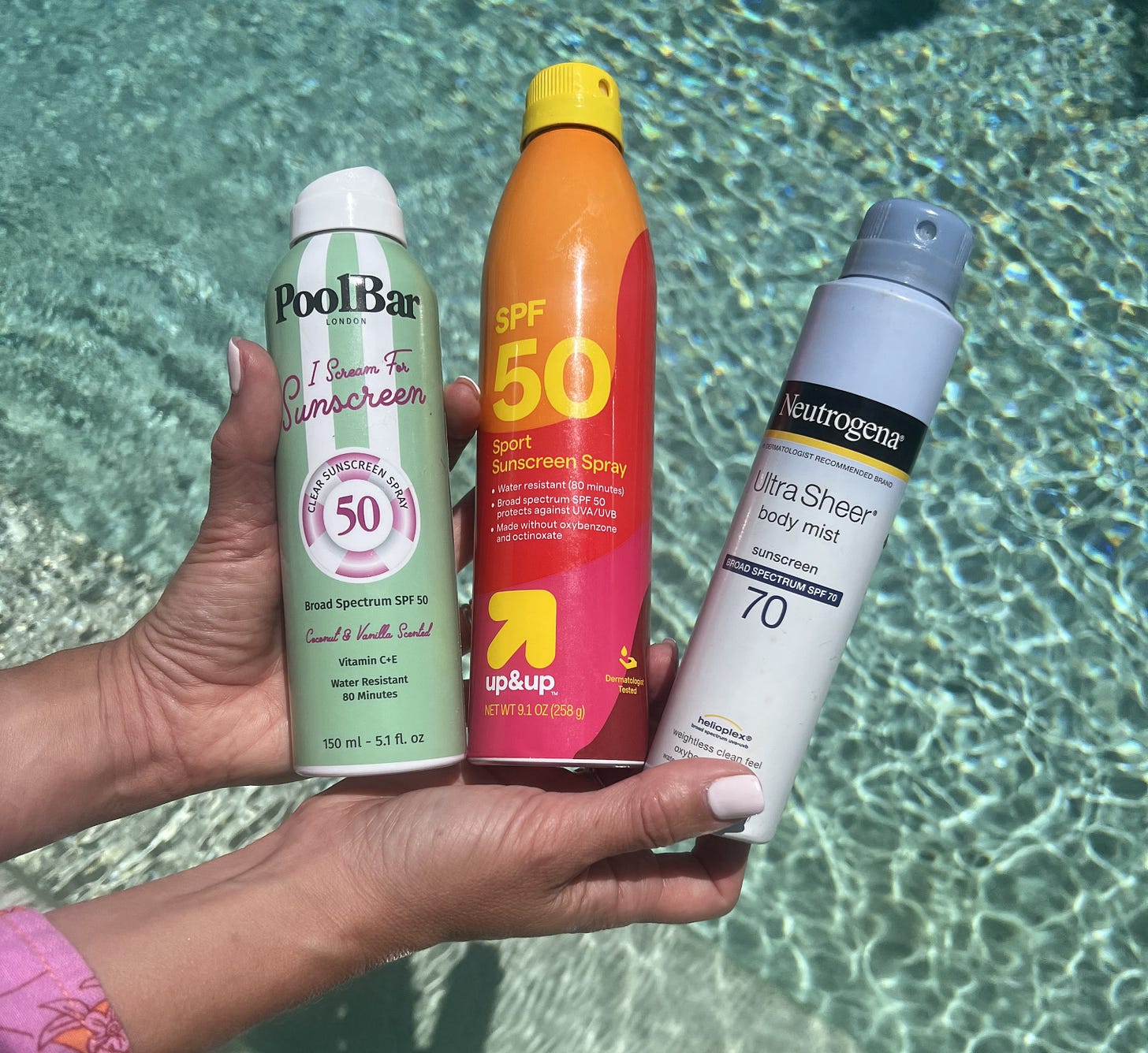 Close-up shot of Beauty Editor Donna Francis holding Pool Bar London I Scream For Sunscreen, Target Up&Up Sport Sunscreen Spray, and Neutrogena Ultra Sheer Body Mist by a shimmering blue swimming pool.
