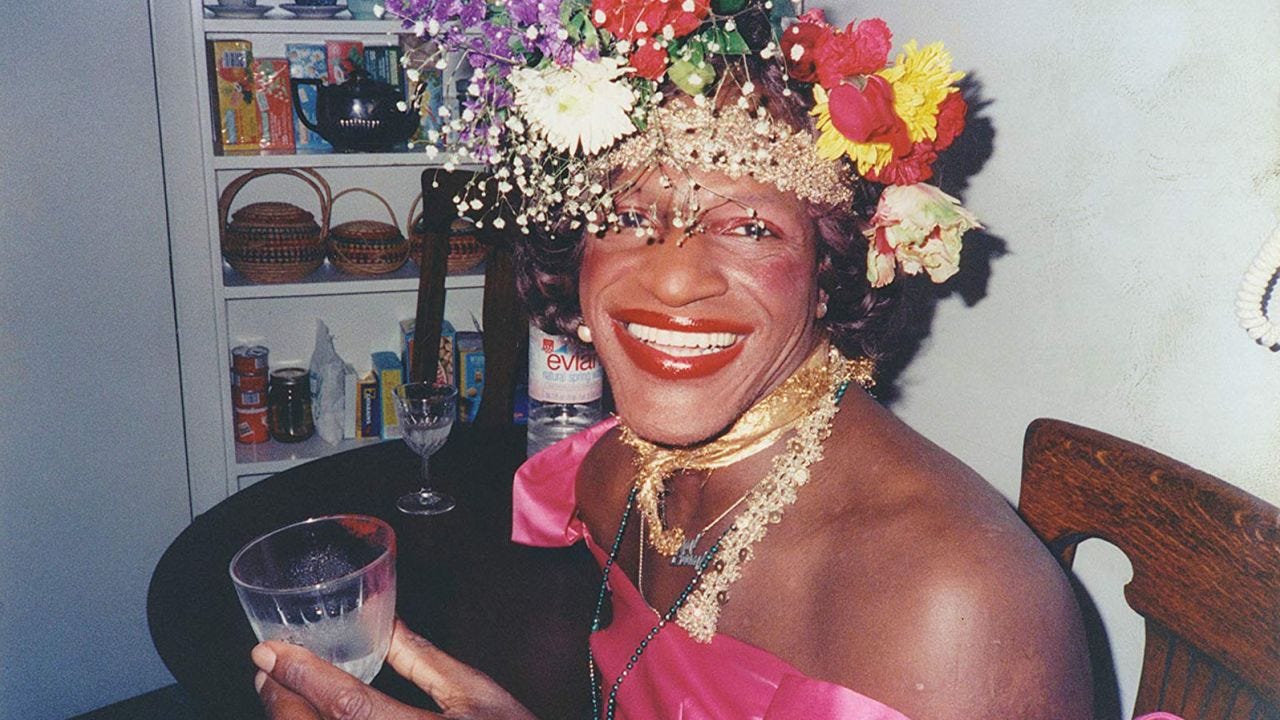 A photograph of Marsha P. Johnson, a black transgender woman. She's wearing a flower crown and bright red lipstick, beaming at the camera.