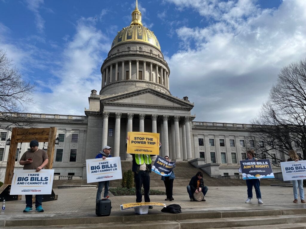 People holding protest signs chant in a row on the steps of a large stone edifice with a gold dome against a partially overcast sky.