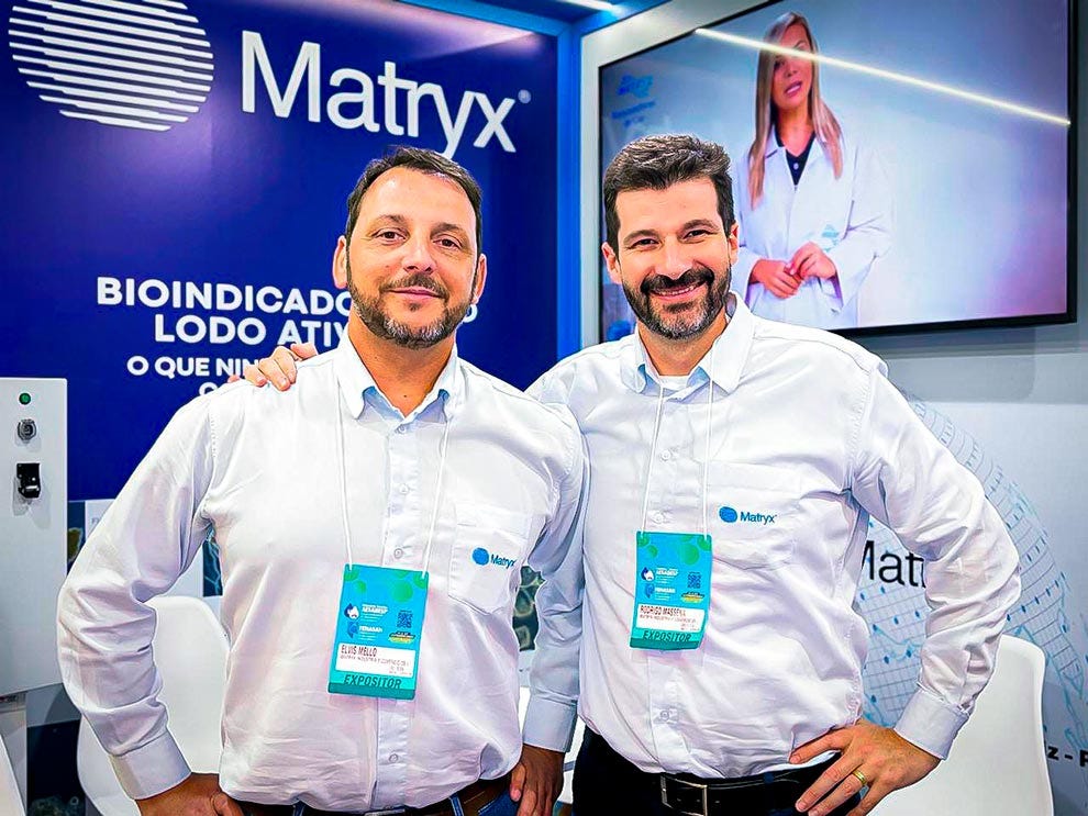 Duas pessoas usando camisas brancas de manga comprida com crachás de identificação azuis e verdes posicionados no peito estão em pé lado a lado em frente a um painel azul com o logotipo e texto "Matryx" e referências a "Bioindicador Lobo Atita", com uma tela exibindo uma profissional em jaleco branco ao fundo, ambiente de evento ou apresentação corporativa. Duas pessoas usando camisas brancas de manga comprida com crachás de identificação azuis e verdes posicionados no peito estão em pé lado a lado em frente a um painel azul com o logotipo e texto "Matryx" e referências a "Bioindicador Lobo Atita", com uma tela exibindo uma profissional em jaleco branco ao fundo, ambiente de evento ou apresentação corporativa.