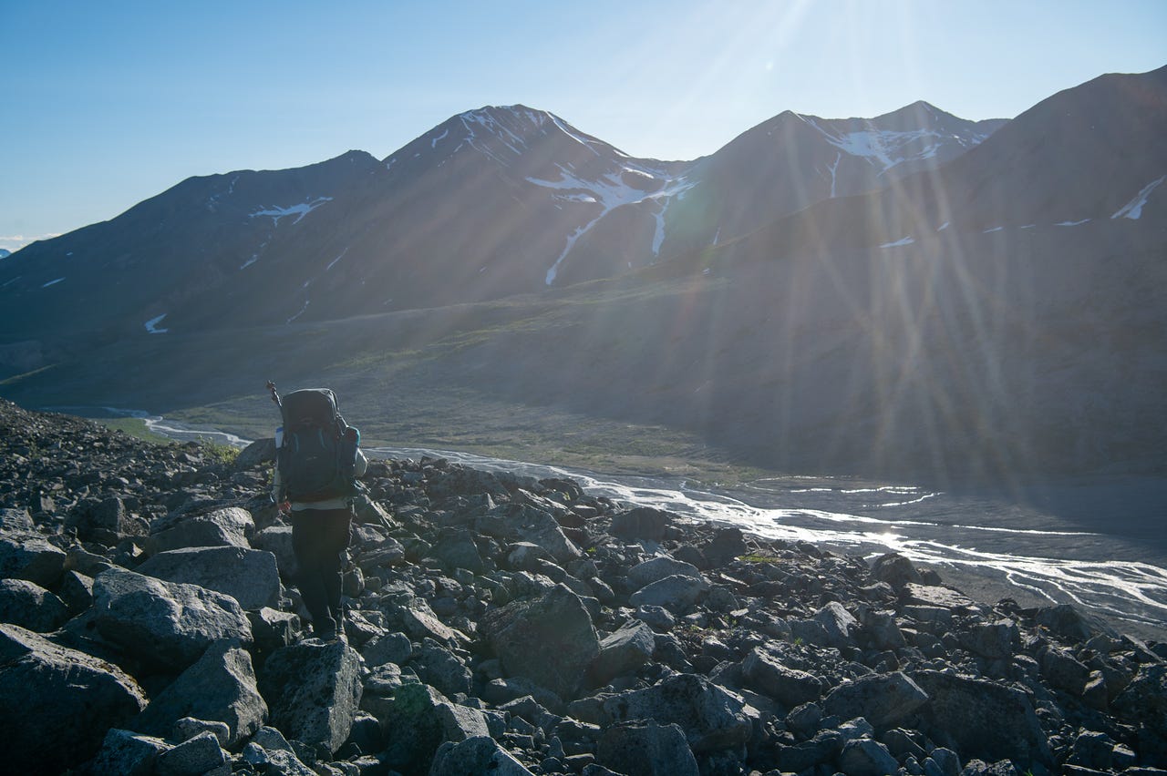 A hiker with a pack picks a way across a jumble of large, dark, unstable boulders on top of a moraine, trekking pole in hand. Beyond, low sun floods a broad braided river bar and silhouettes the mountains ringing the valley, sunbeams slanting across the frame.