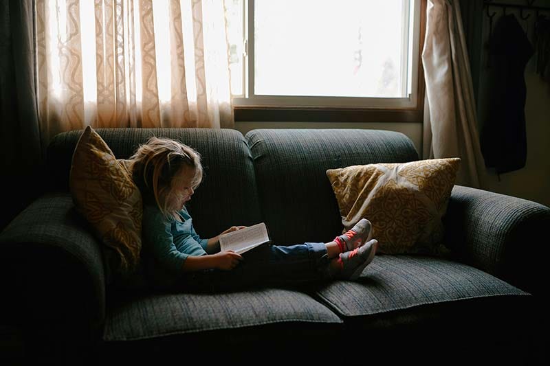 Une petite fille lit un livre, assise dans un canapé gris, près d'une fenêtre.