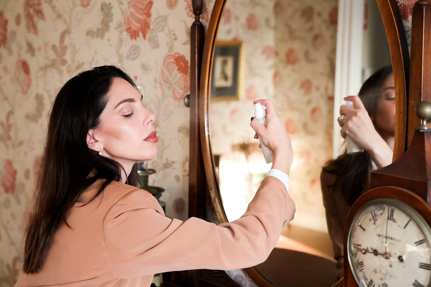 A close-up of a woman with long dark hair and dewy skin wearing a peach-colored outfit. She sprays a bottle of mist near her face while looking serene. A vintage mirror and wallpaper with a floral pattern create a classic, elegant setting in the background.
