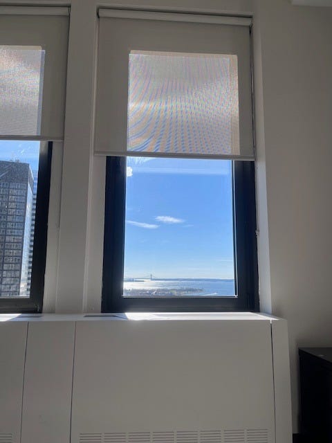 View of blue sky, one skyscraper, and the horizon of NY Harbor our of a white institutional window frame, its shade half-closed. View of blue sky, one skyscraper, and the horizon of NY Harbor our of a white institutional window frame, its shade half-closed.